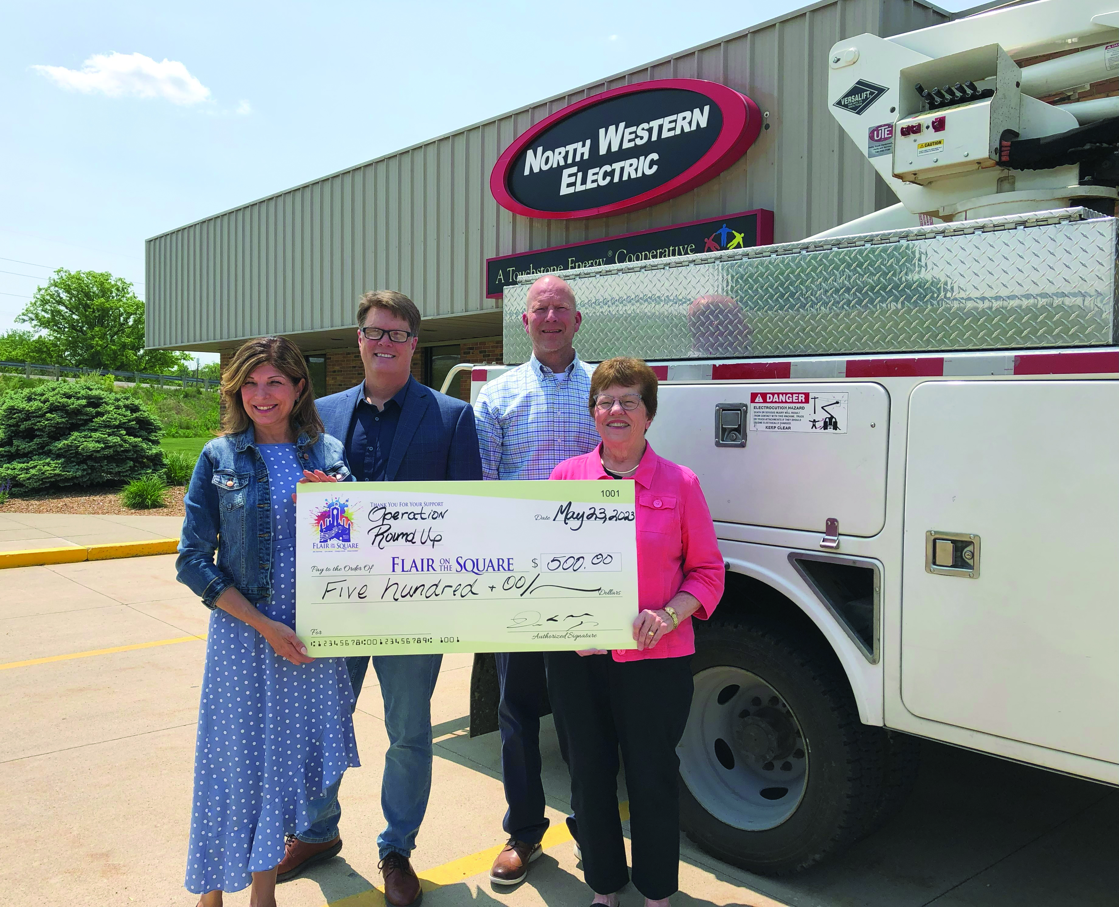 FOUR ADULTS HOLDING BIG CHECK AND STANDING IN FRONT OF BUCKET TRUCK