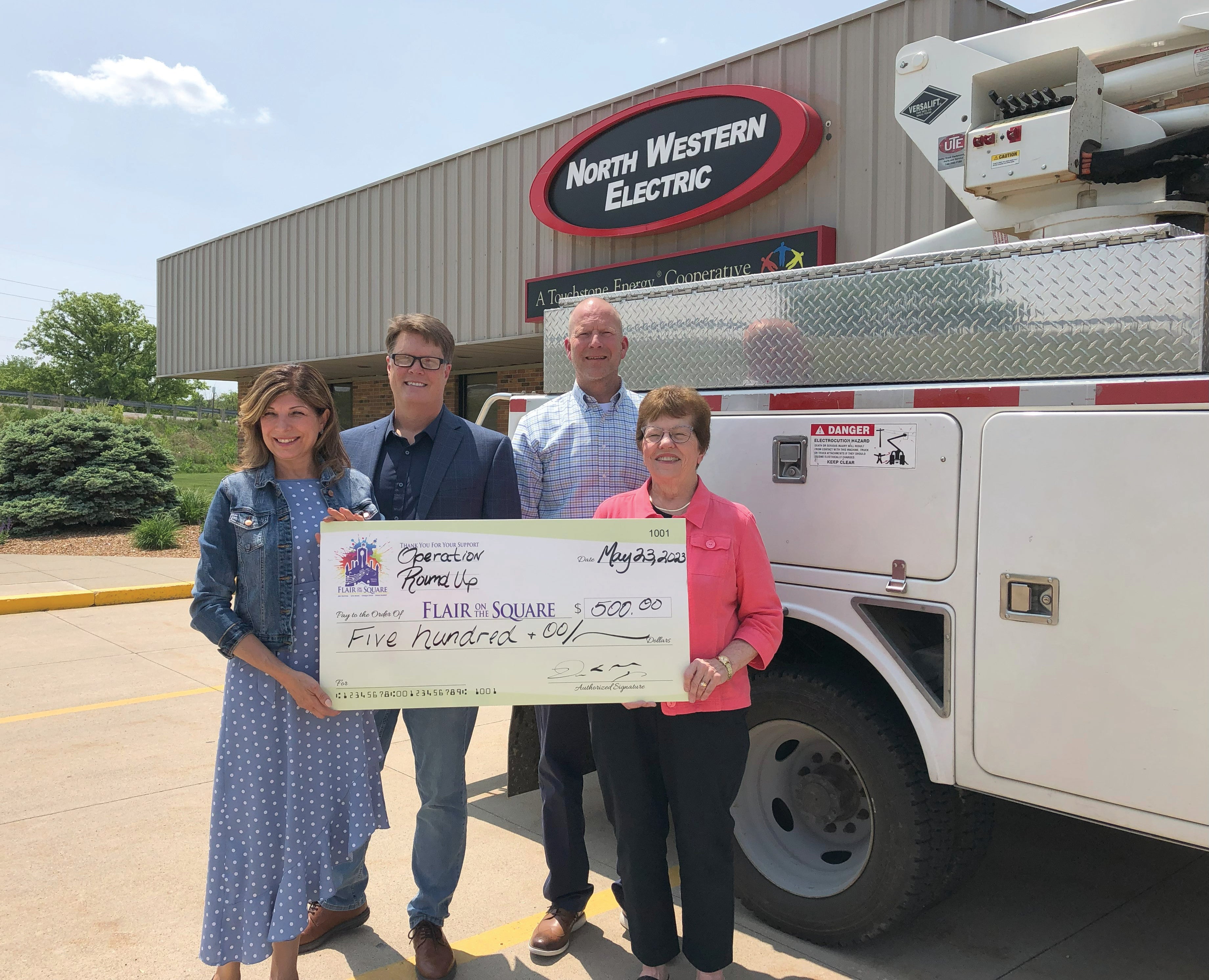FOUR ADULTS HOLDING BIG CHECK AND STANDING IN FRONT OF BUCKET TRUCK